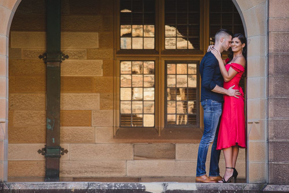 Two people in formal attire pose together against a sandstone building backdrop with arched windows.