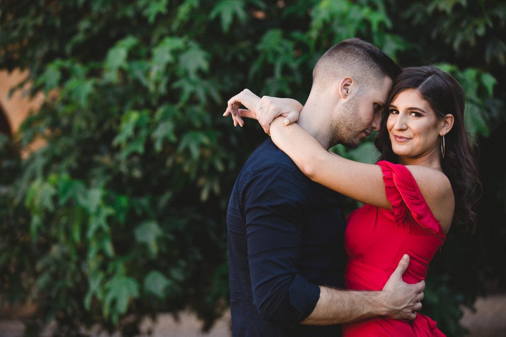 Two people embrace near green foliage against a stone building with dramatic lighting.