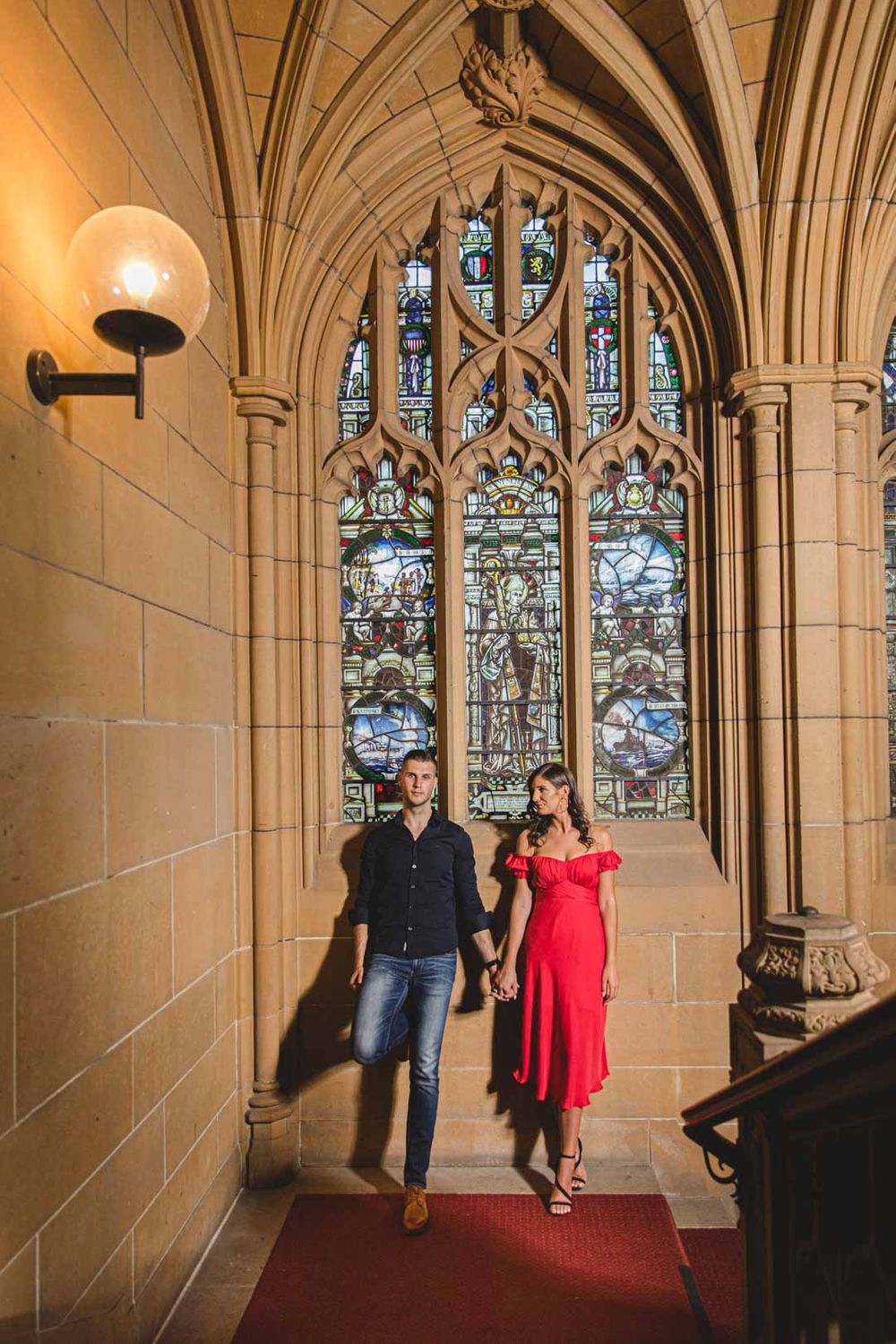 Two people pose in an ornate Gothic corridor with beautiful stained glass windows and vaulted ceilings.