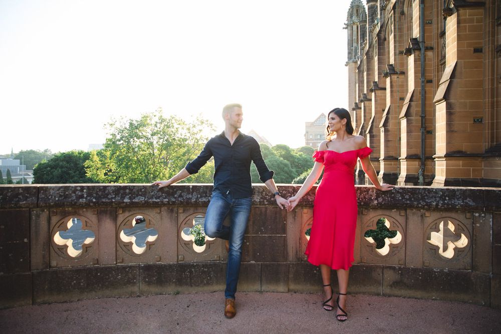 A couple poses along a stone balcony with gothic architectural details at sunset.