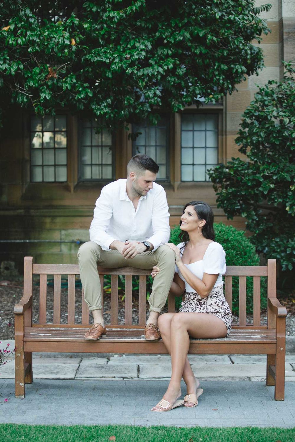 A couple shares a relaxed moment sitting together on a wooden bench surrounded by greenery.