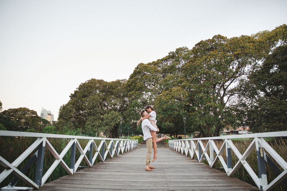 A romantic moment captured on a white wooden bridge surrounded by lush green trees.