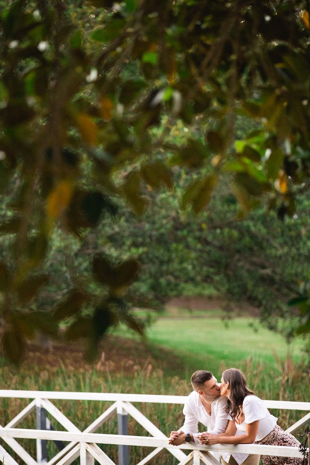A couple sharing a romantic moment on a white railing bridge framed by leafy branches.