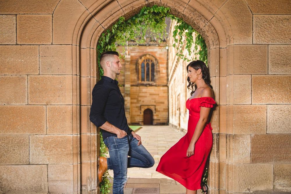 Romantic couple poses together under stone archways at historic building with red dress and dark clothing.
