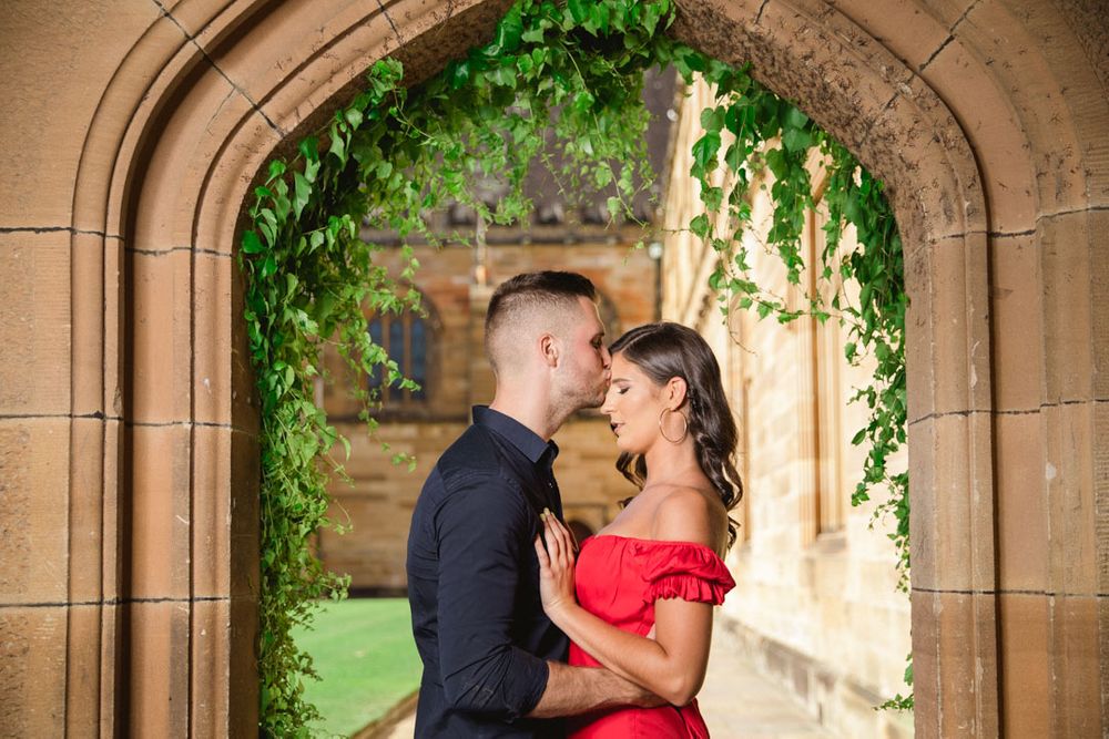 Elegant couple embraces beneath ornate stone arches with green ivy and natural lighting.