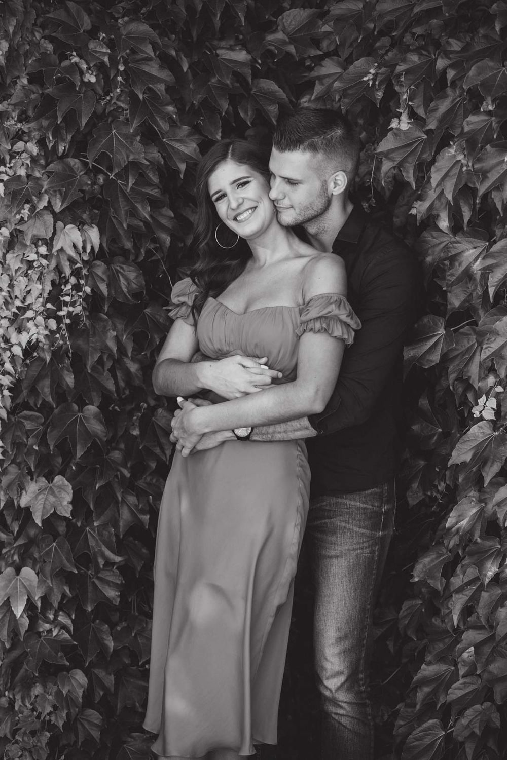 Black and white intimate portrait against ivy-covered wall showing tender embrace.