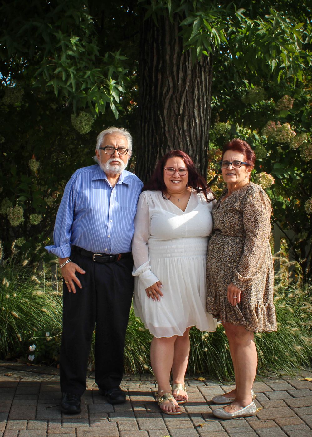 Three people smile together on brick patio with blooming garden and large tree in background.