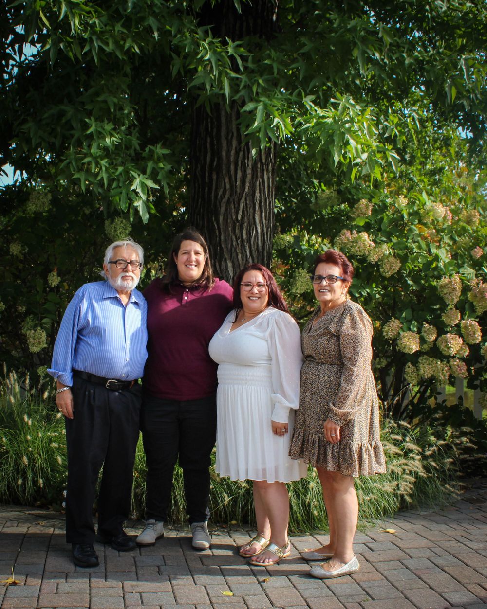 Four people pose together on brick path with large tree and flowering bushes in background.