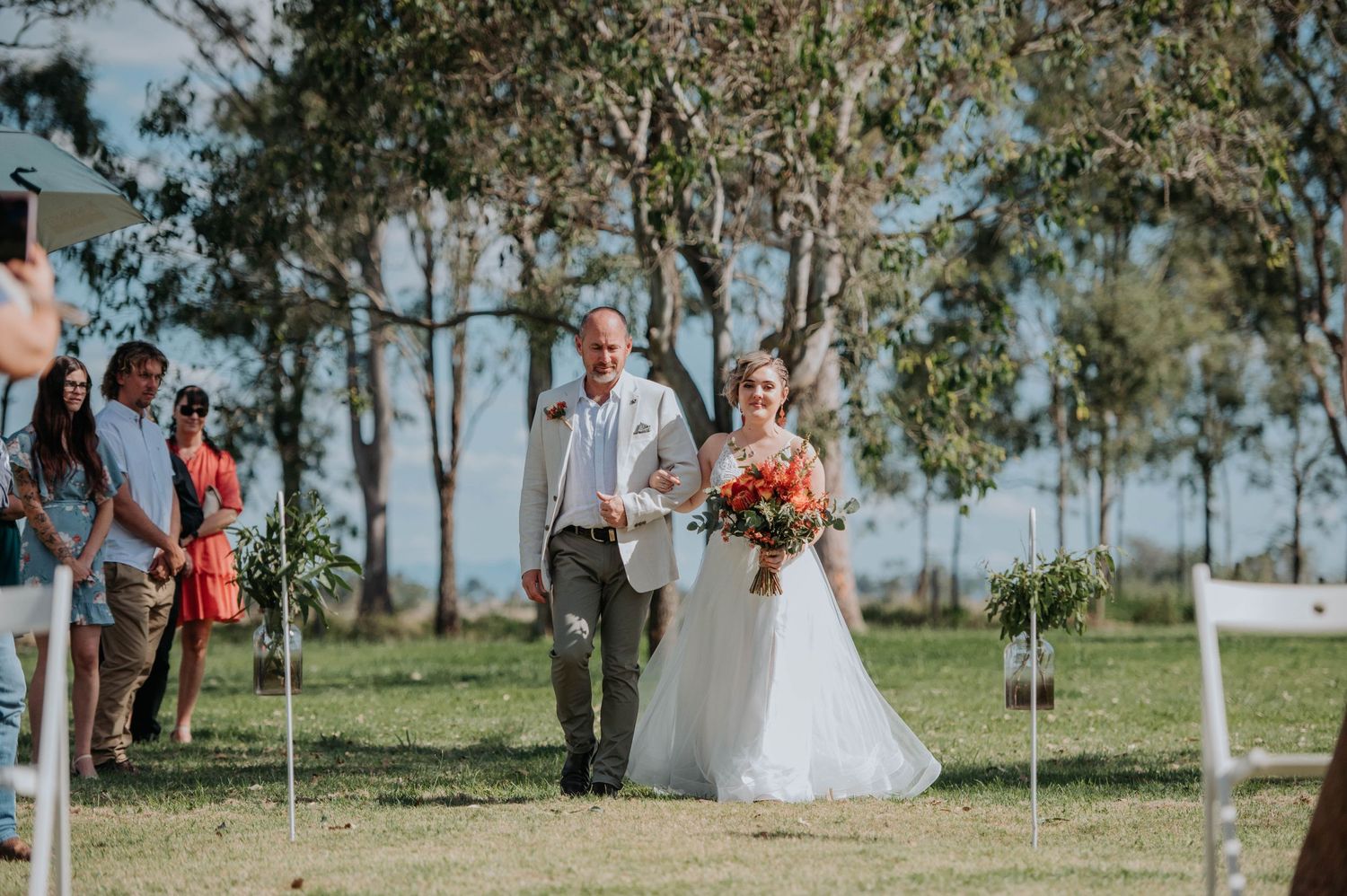 A bride in white dress walks down an outdoor aisle with eucalyptus trees in background.