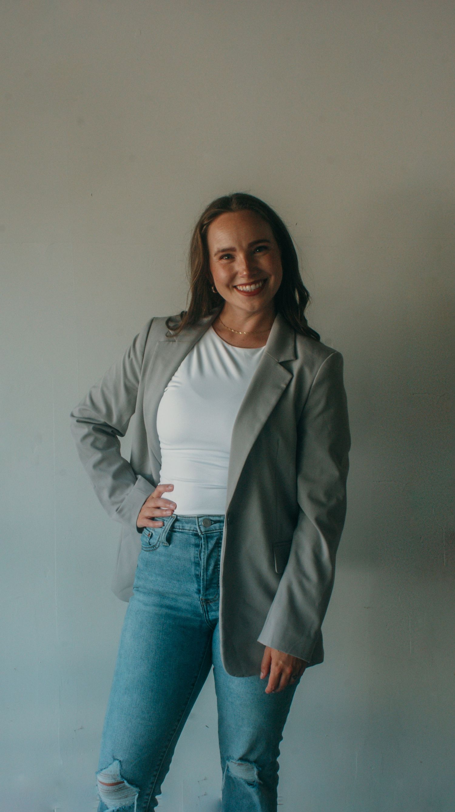 Casual business style portrait in grey blazer and jeans against neutral wall.