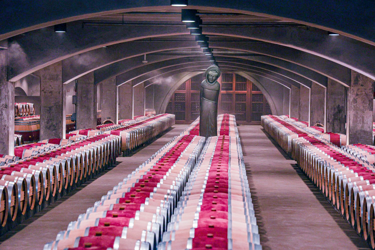 Long wine cellar with arched ceiling and rows of barrels lined with red and white fabric.