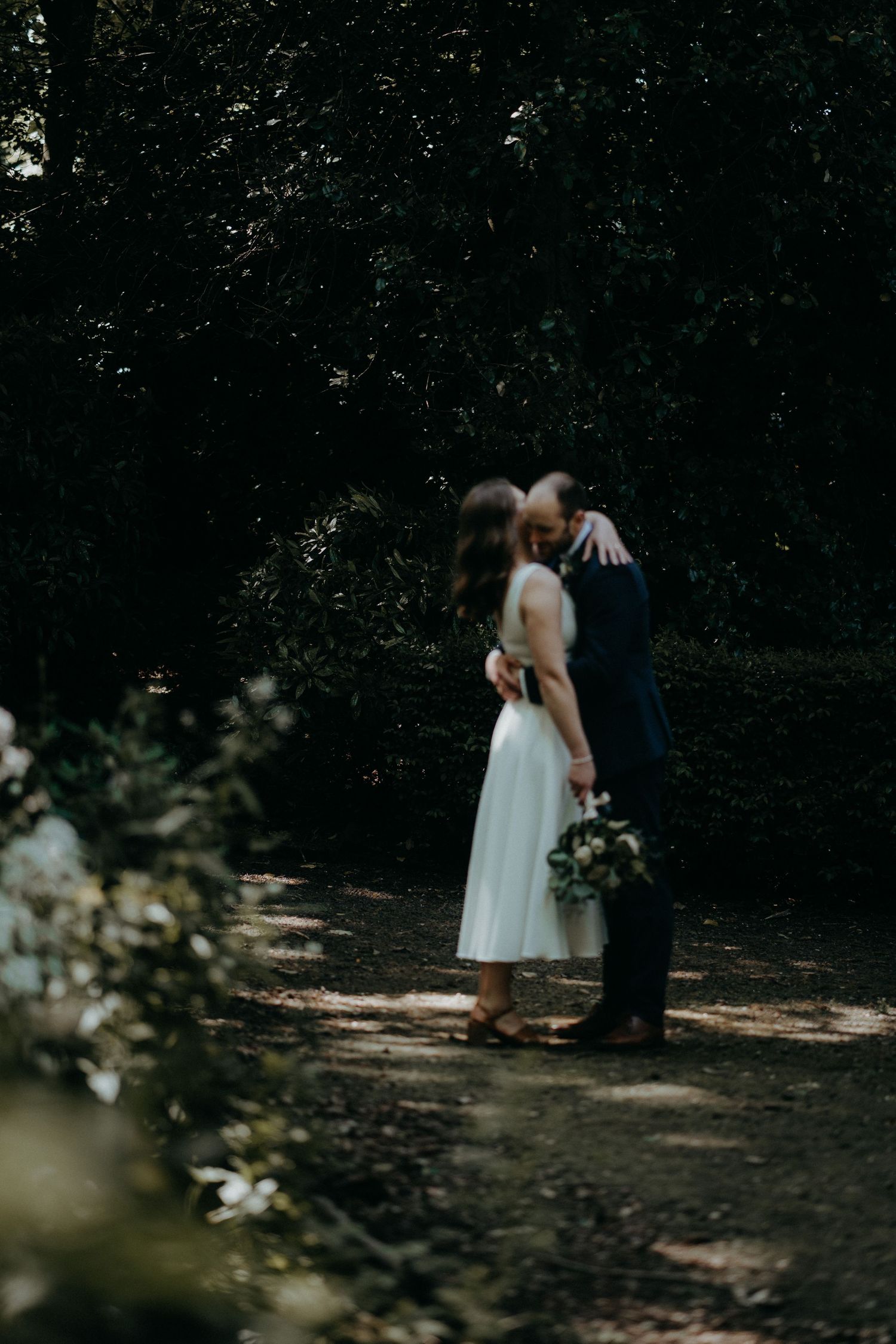 A romantic wedding couple stands together on a wooded path in soft lighting.