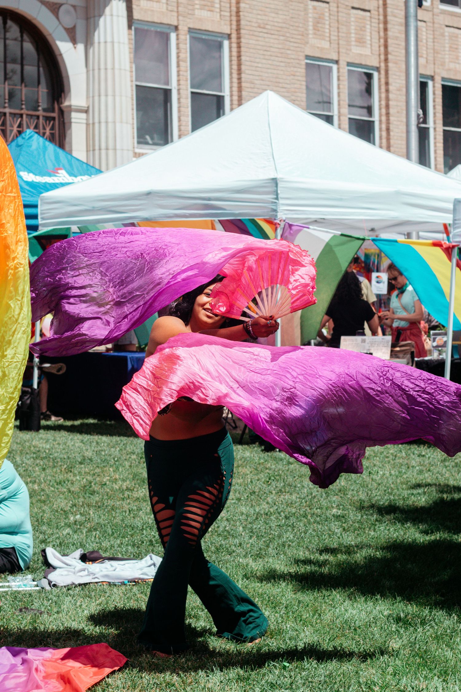 Performer with colorful silk fans dances at outdoor festival under white tent.
