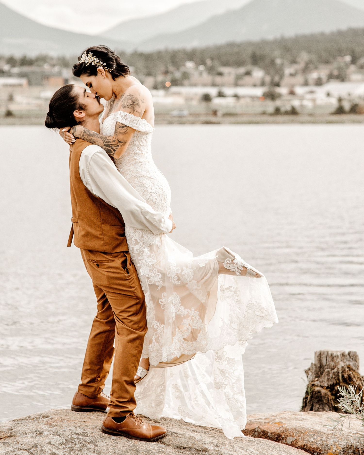 A romantic wedding couple embraces on rocky shores beside a mountain lake with lush scenery in the background.