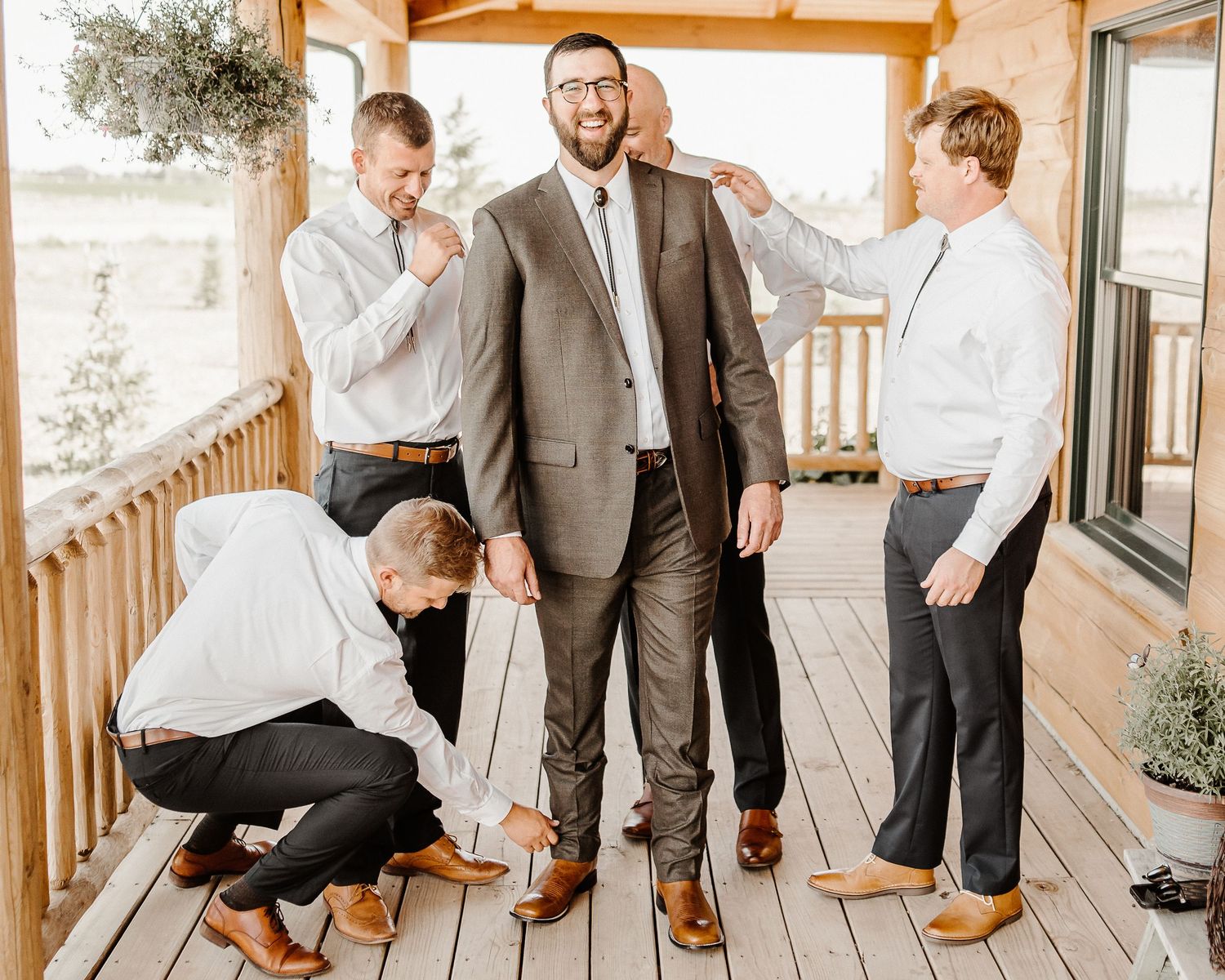 Groomsmen share a candid moment on a wooden porch, laughing together while adjusting formal attire before wedding.