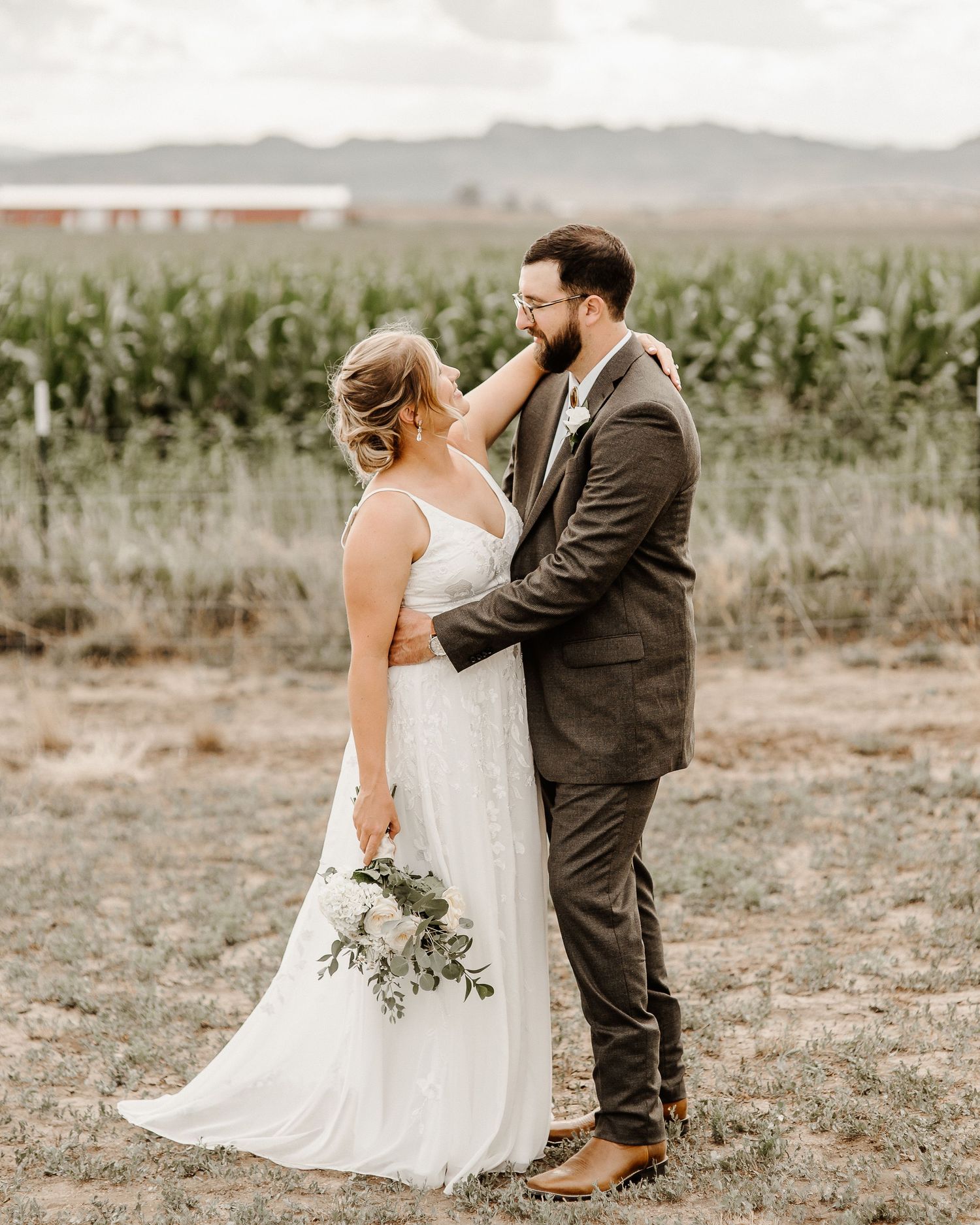 A couple in wedding attire stands in front of a corn field on their special day in a rustic outdoor setting.