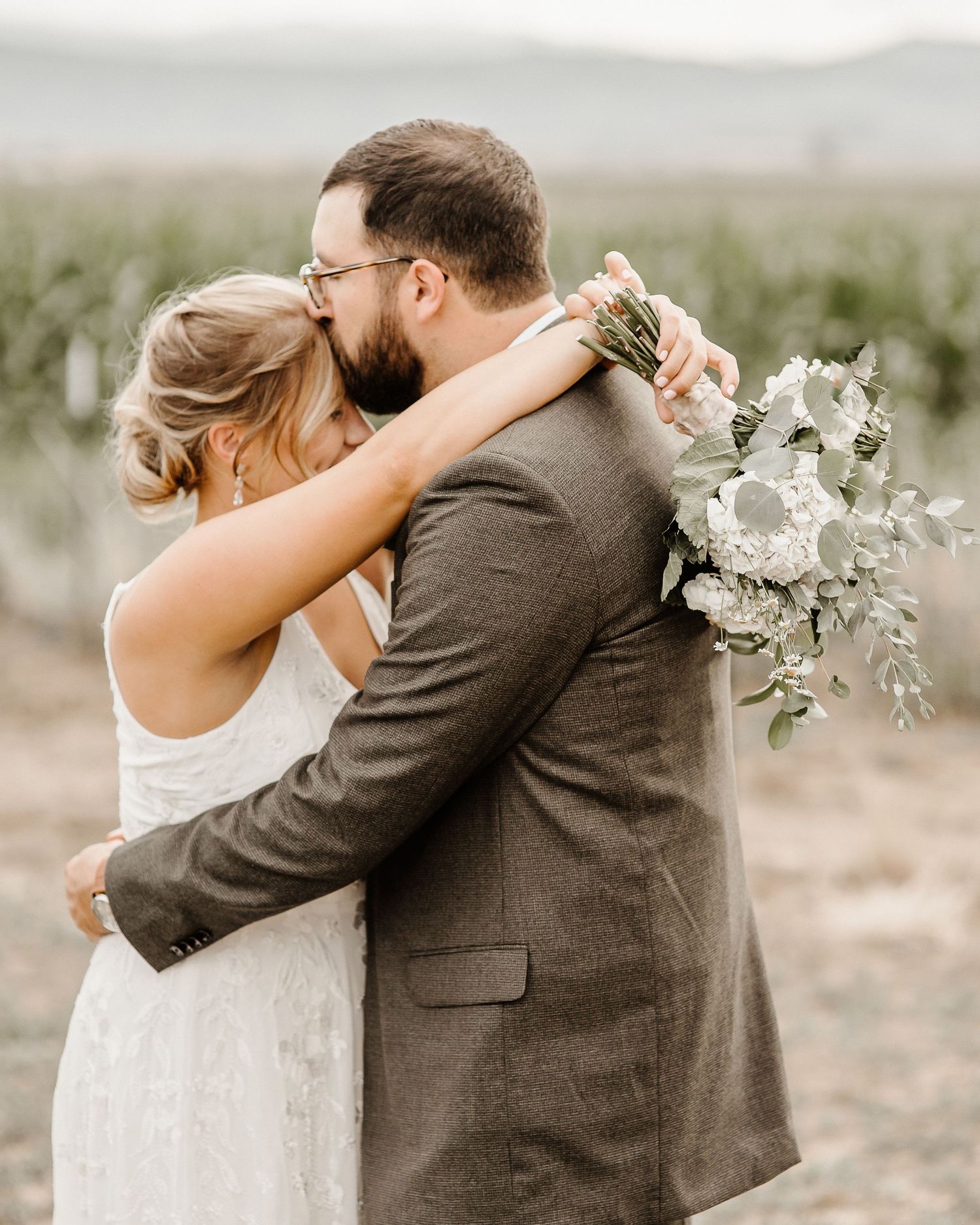 A couple in wedding attire embrace outdoors, with the bride holding a white and green eucalyptus bouquet.