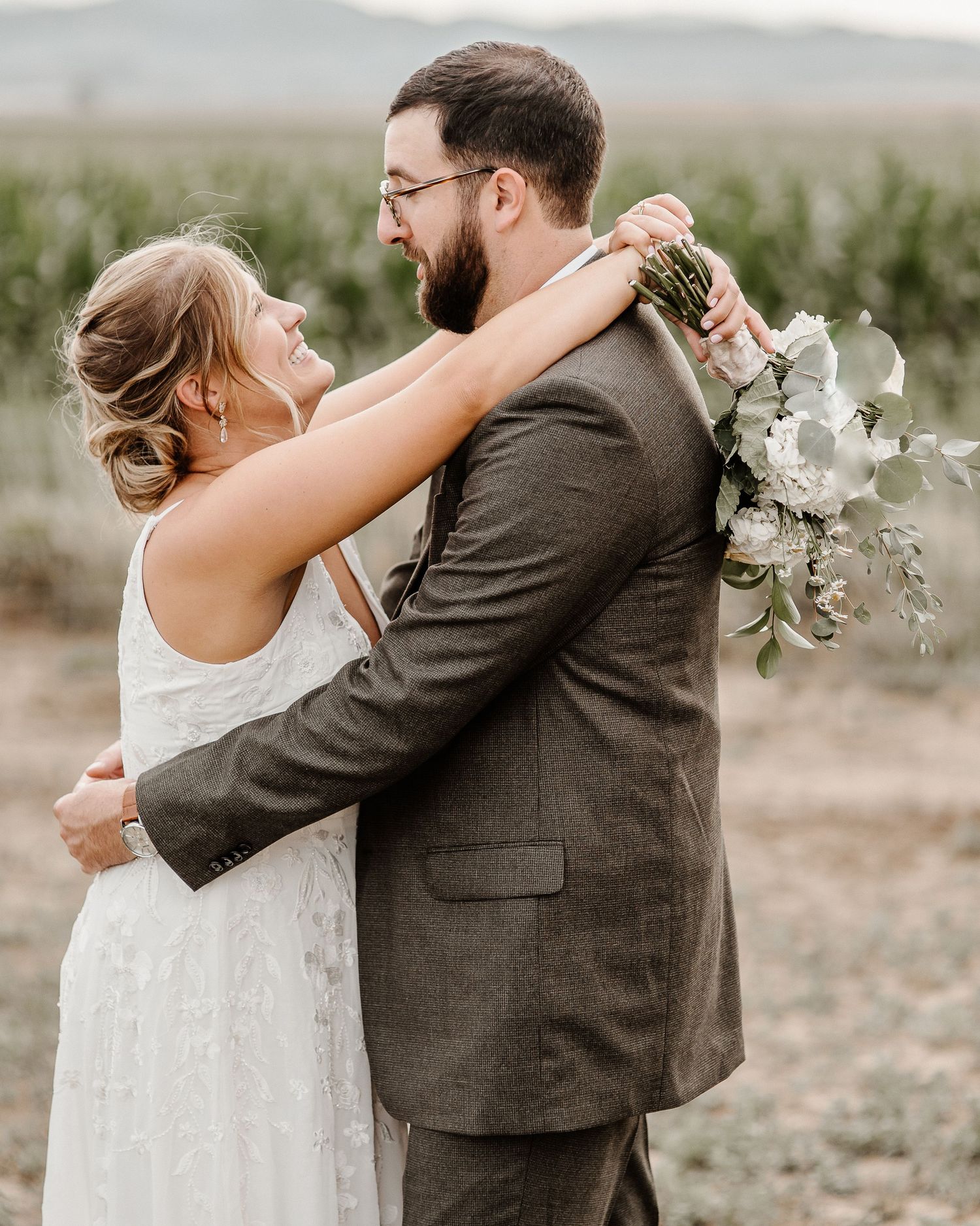 A couple dressed in wedding attire share an intimate embrace outdoors while holding a white floral bouquet.