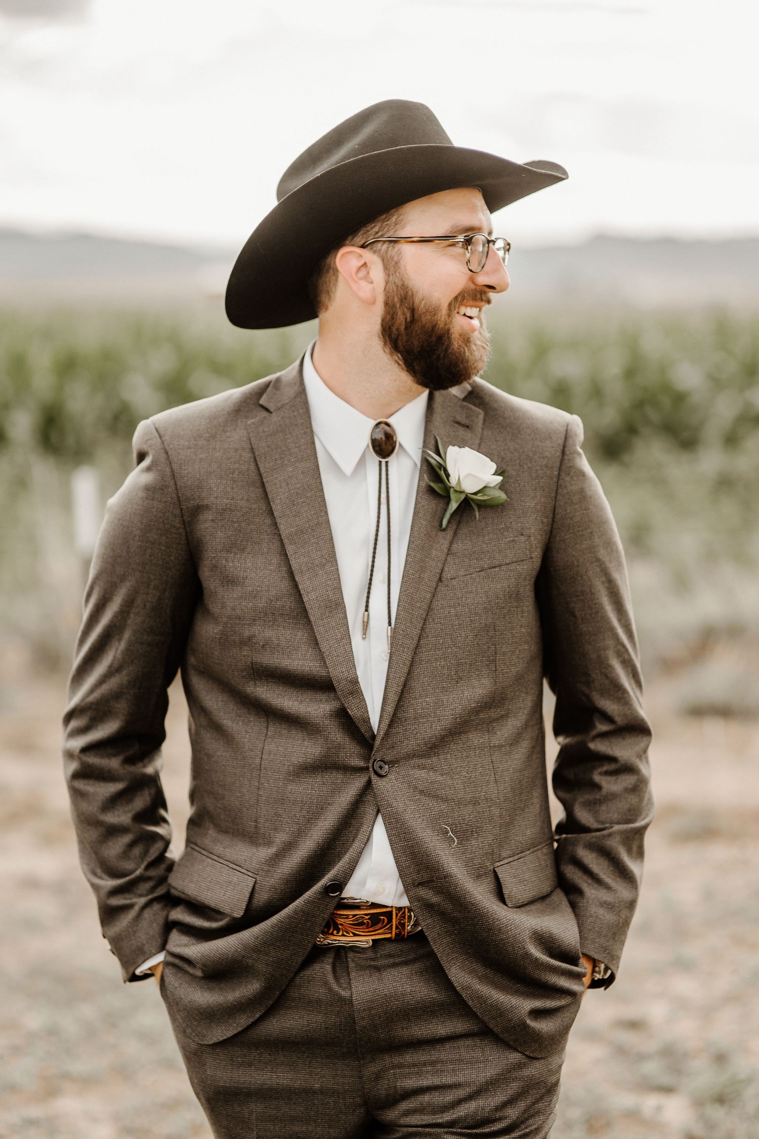 A groom in a brown suit and cowboy hat smiles while standing outdoors on his western-themed wedding day.