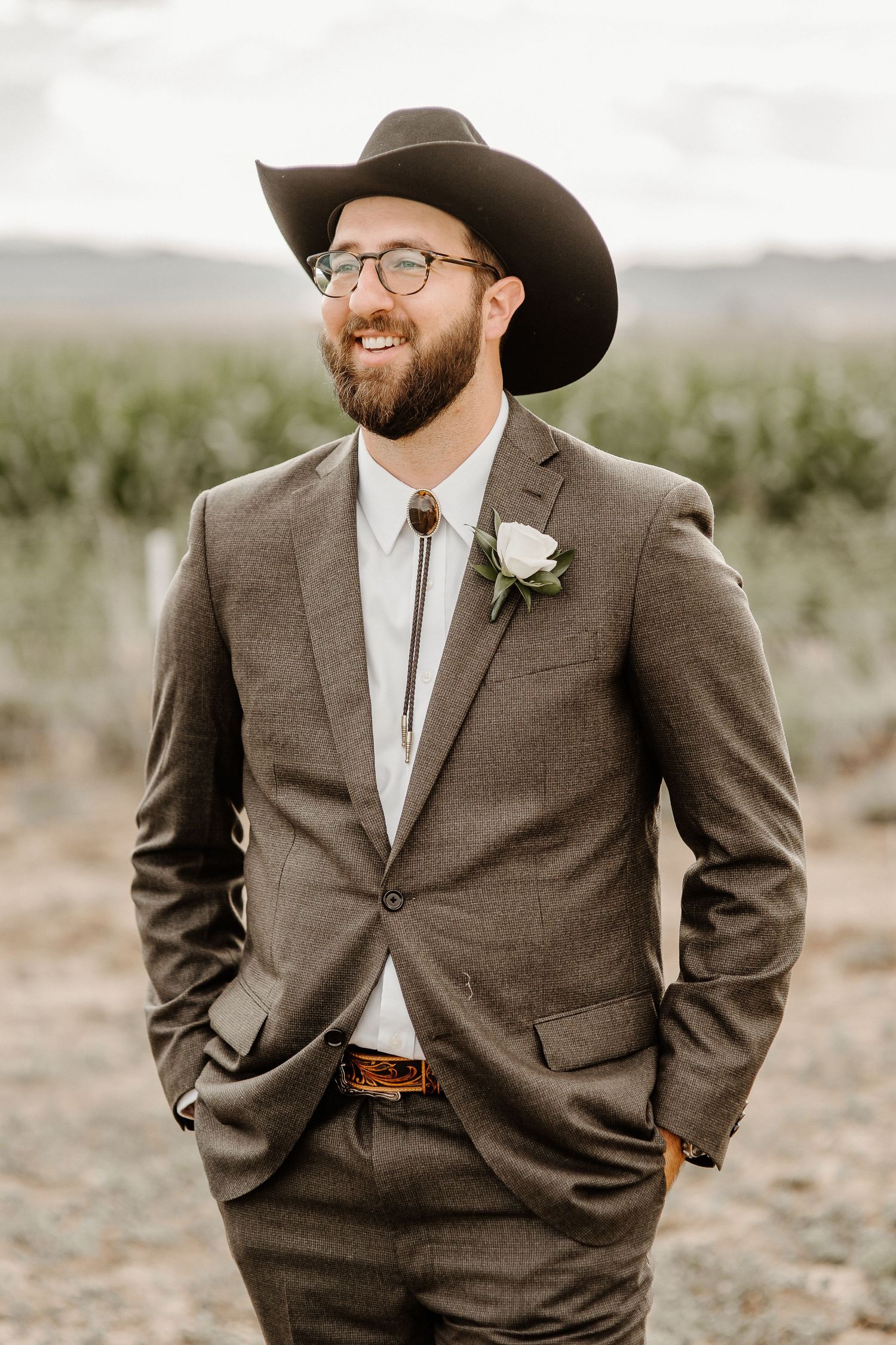 A groom in a brown suit, cowboy hat and glasses smiles while wearing a white boutonniere against a desert landscape.