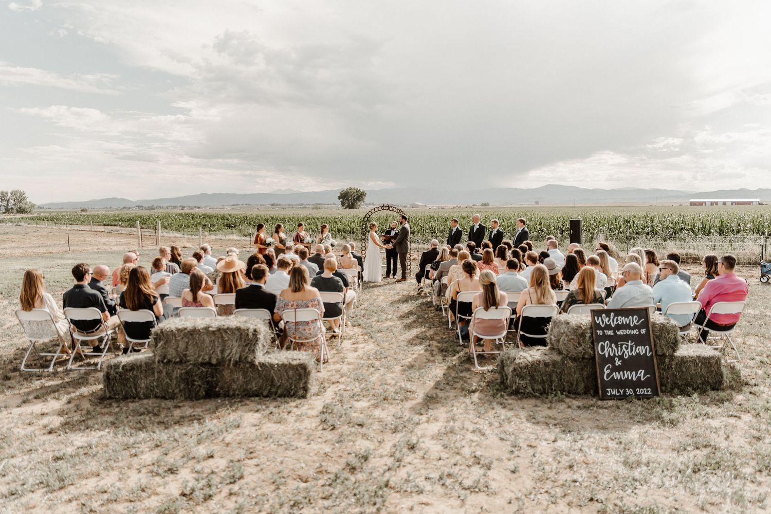 A rustic outdoor wedding ceremony with hay bale seating surrounded by farm fields under a cloudy sky.