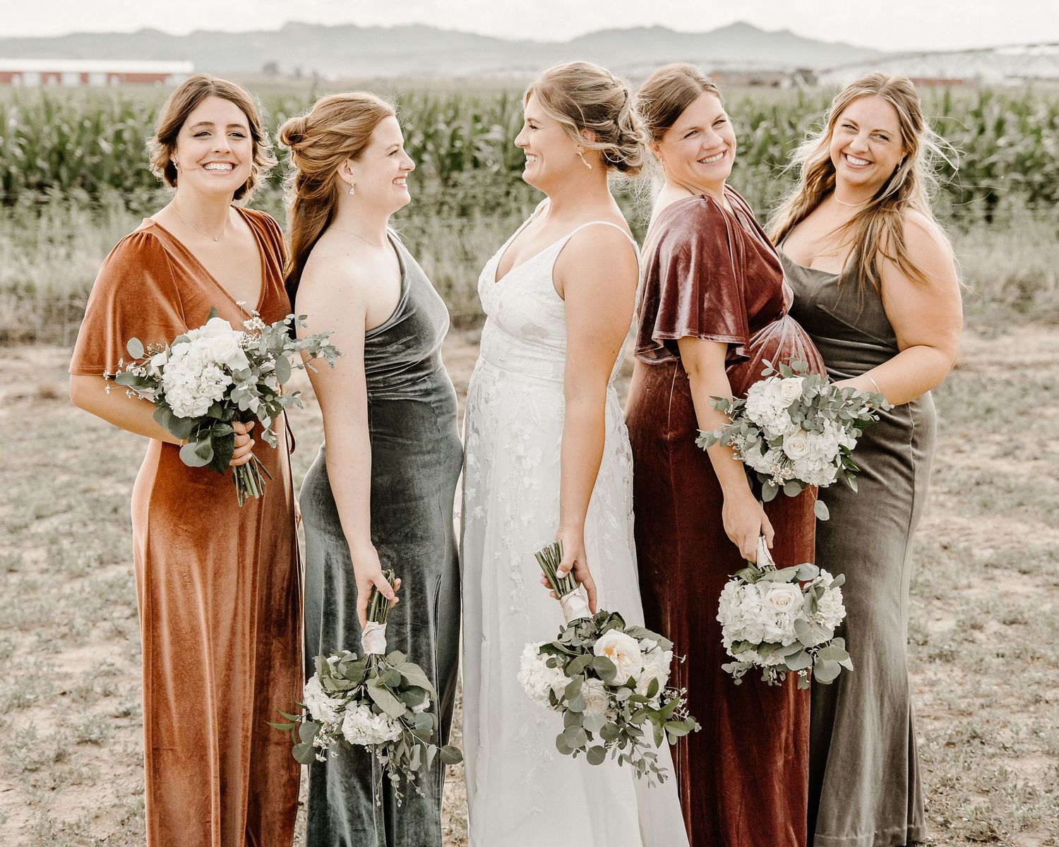 A bridal party in velvet mix-and-match dresses holds white floral bouquets while standing in a misty outdoor field.