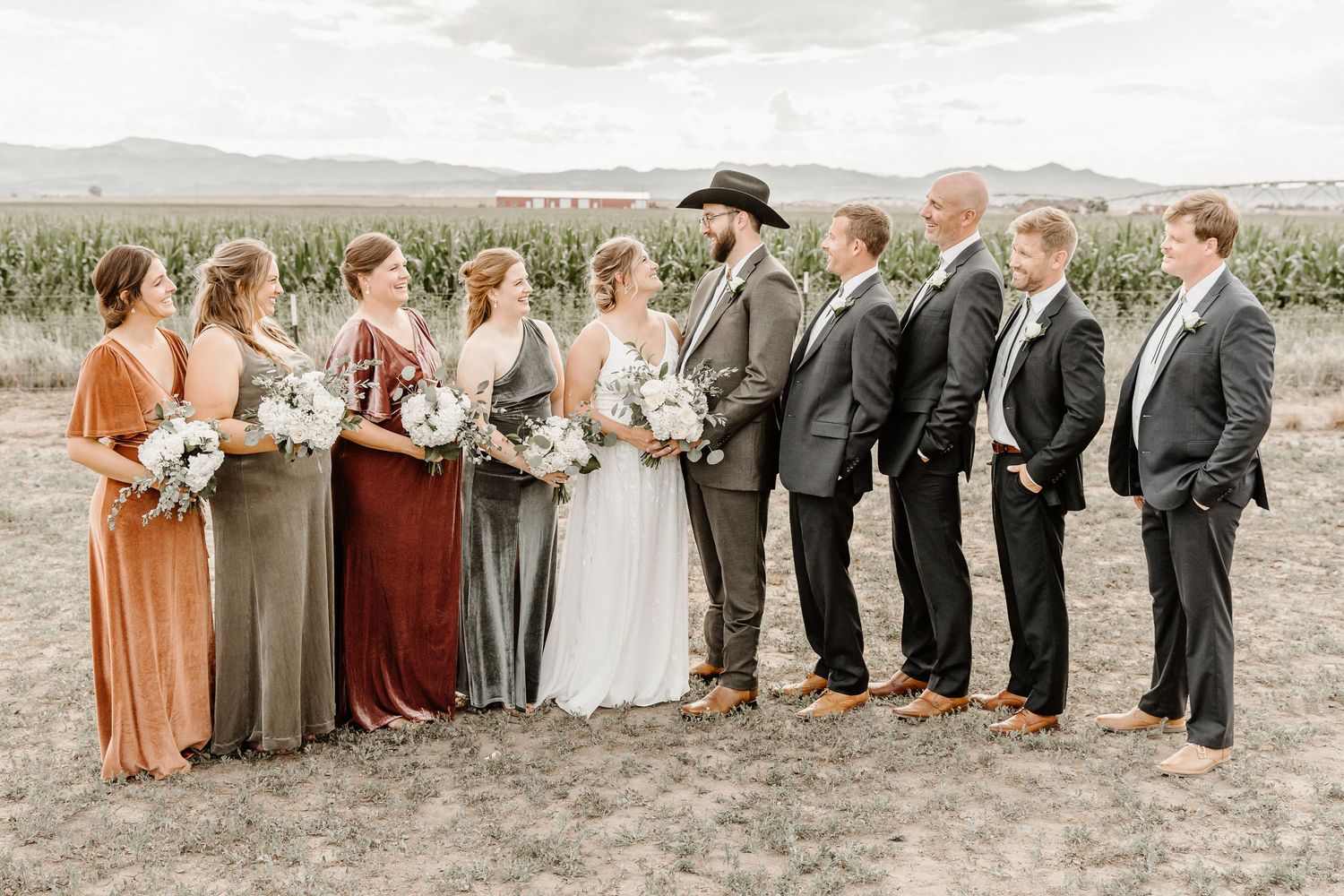 Wedding party in an outdoor rustic setting with bridesmaids in earth-toned dresses and groomsmen in dark suits.