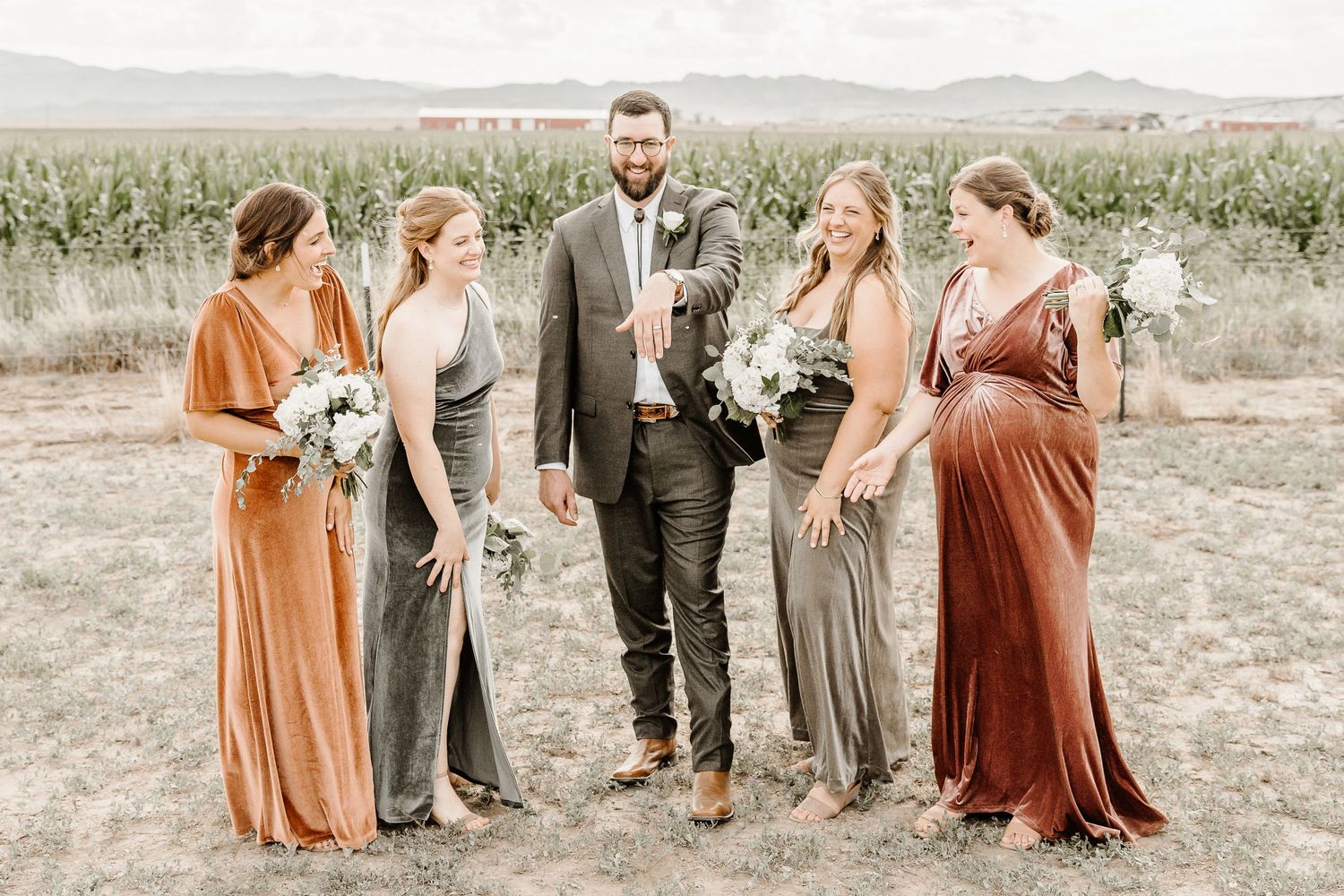 Wedding party in earth-toned dresses and suits pose together in an open field with mountains in background.