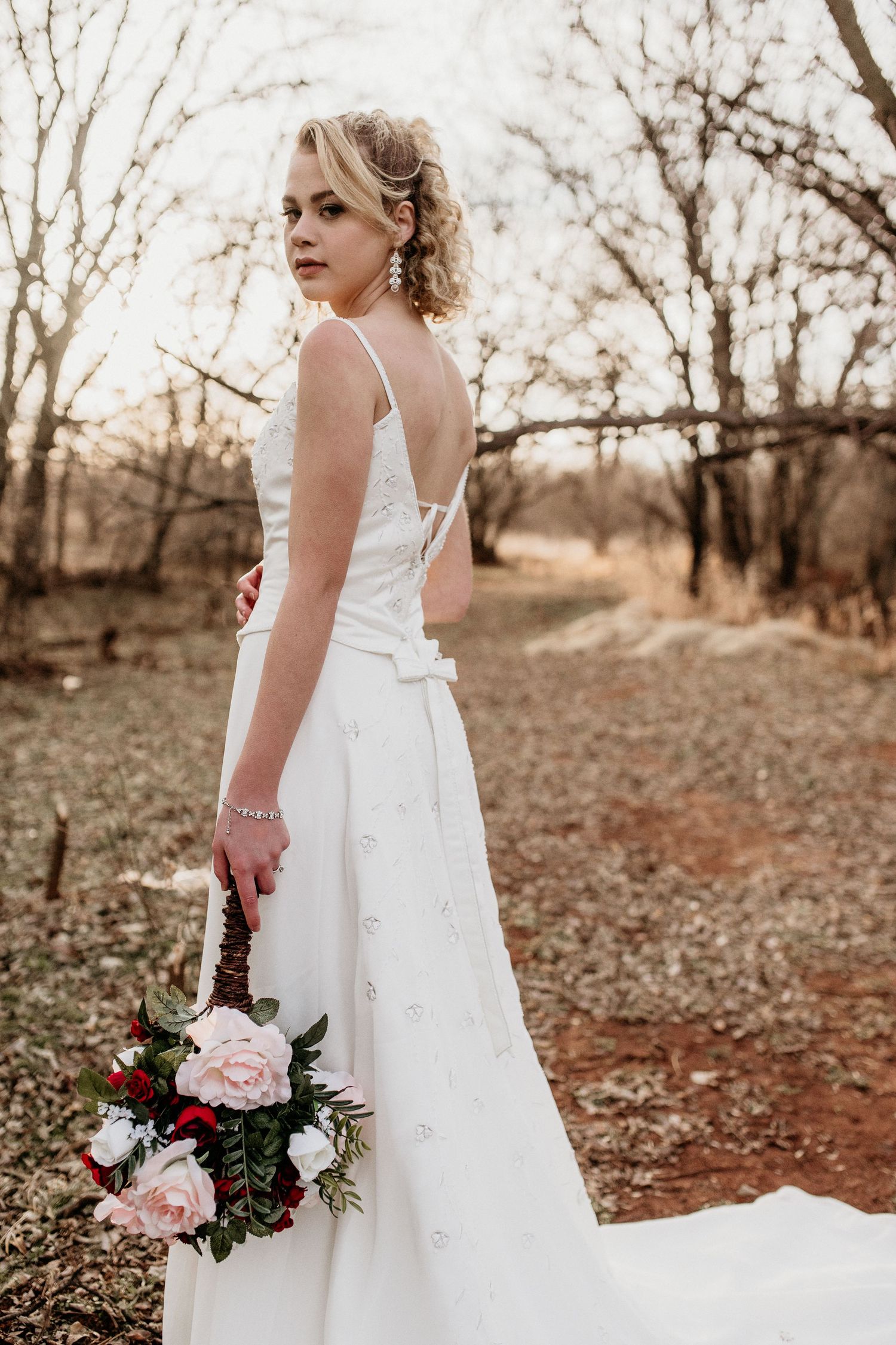 A bride in a flowing white wedding gown holds a pink and burgundy bouquet while standing on a rustic autumn path.