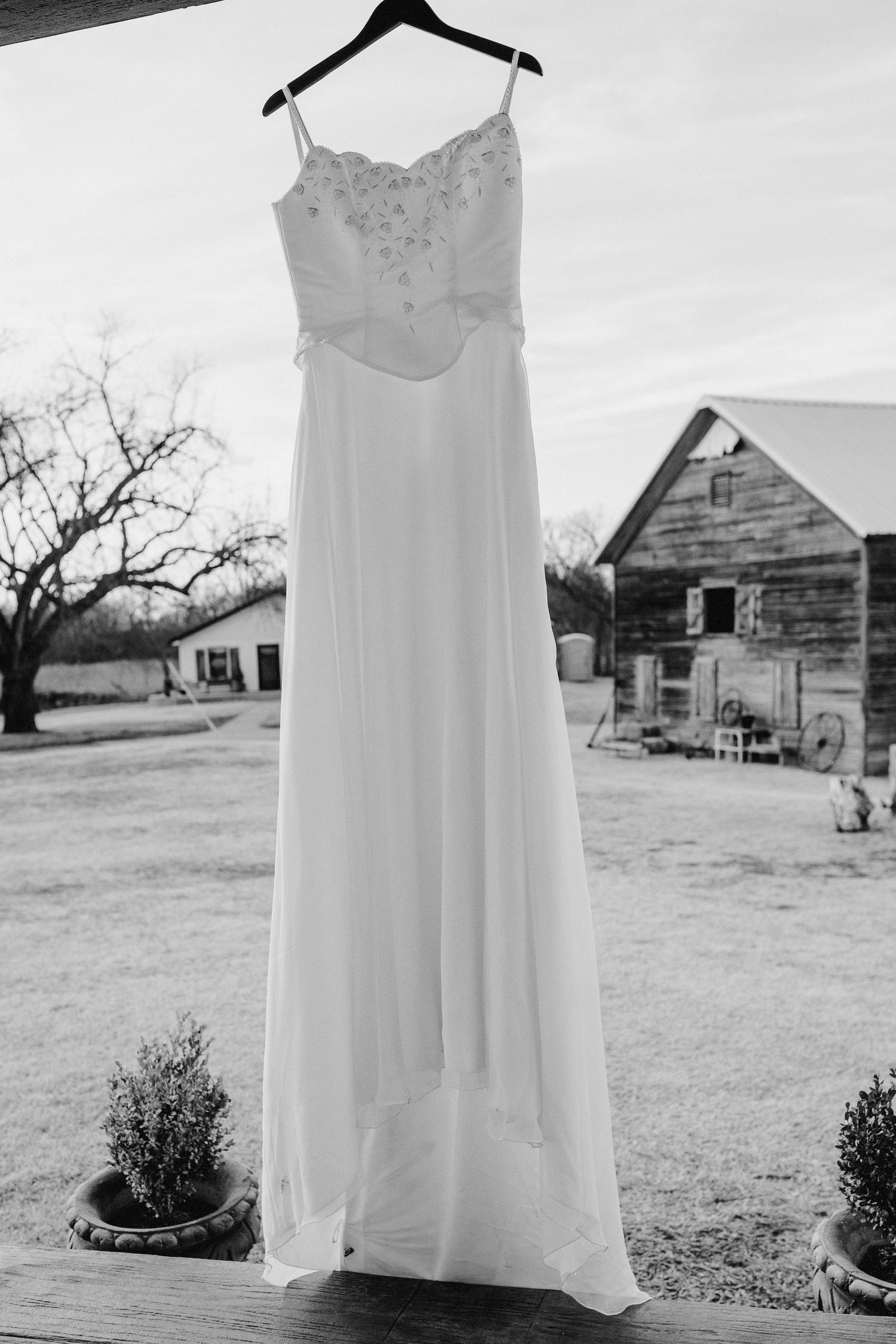 A white wedding dress with spaghetti straps hangs on display against a rustic barn backdrop in black and white.