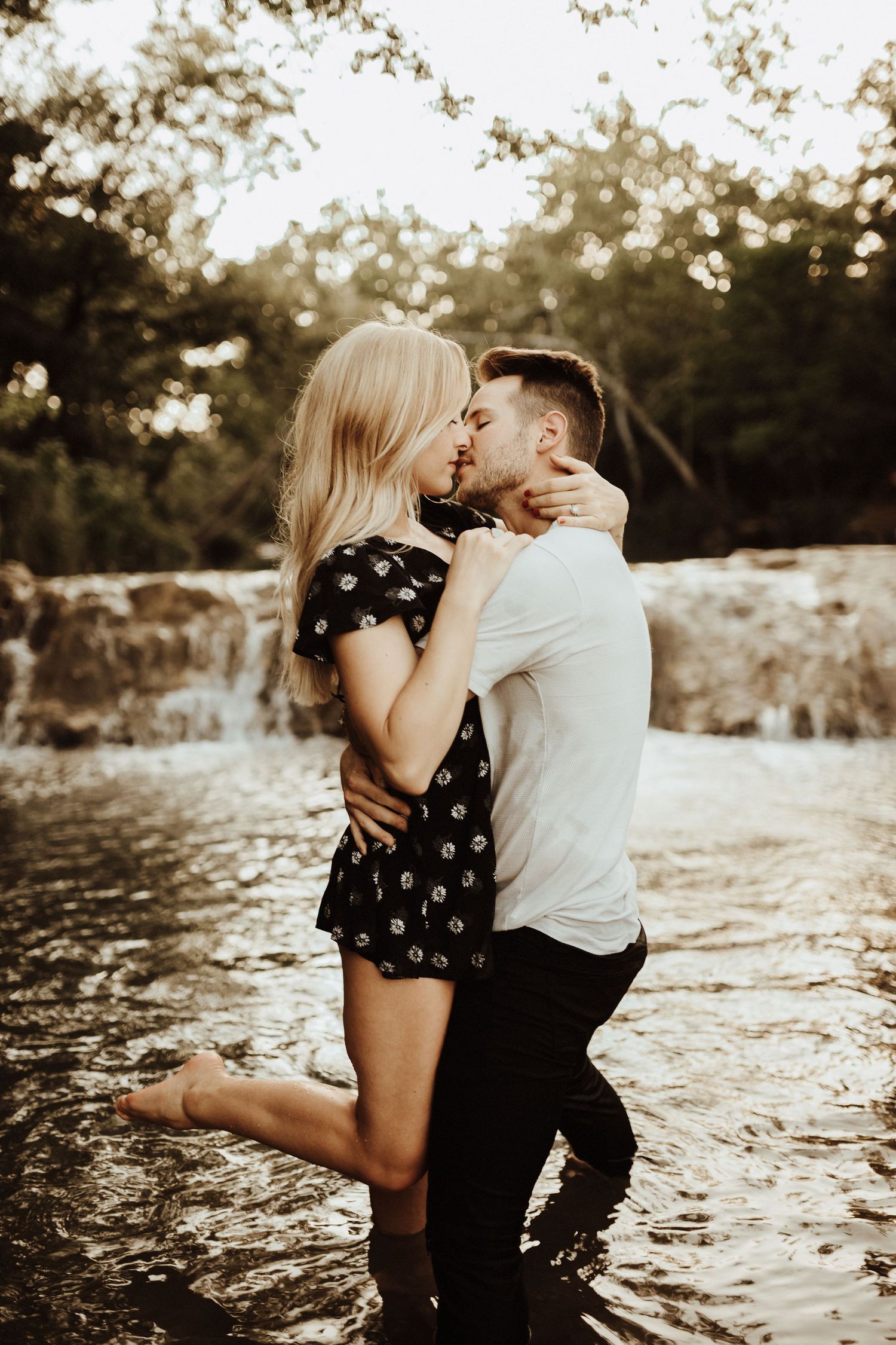 Romantic couple sharing an intimate moment by a waterfall at sunset in a moody, warm-toned setting.