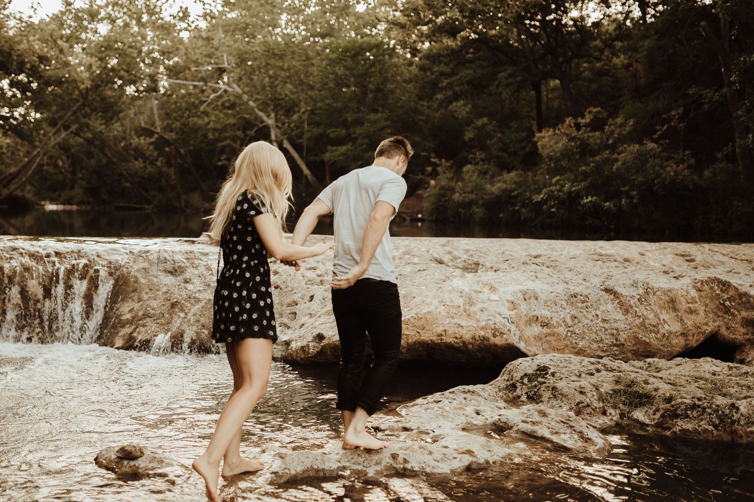 Two people hold hands while exploring rocky terrain near a waterfall on a sunny summer day.