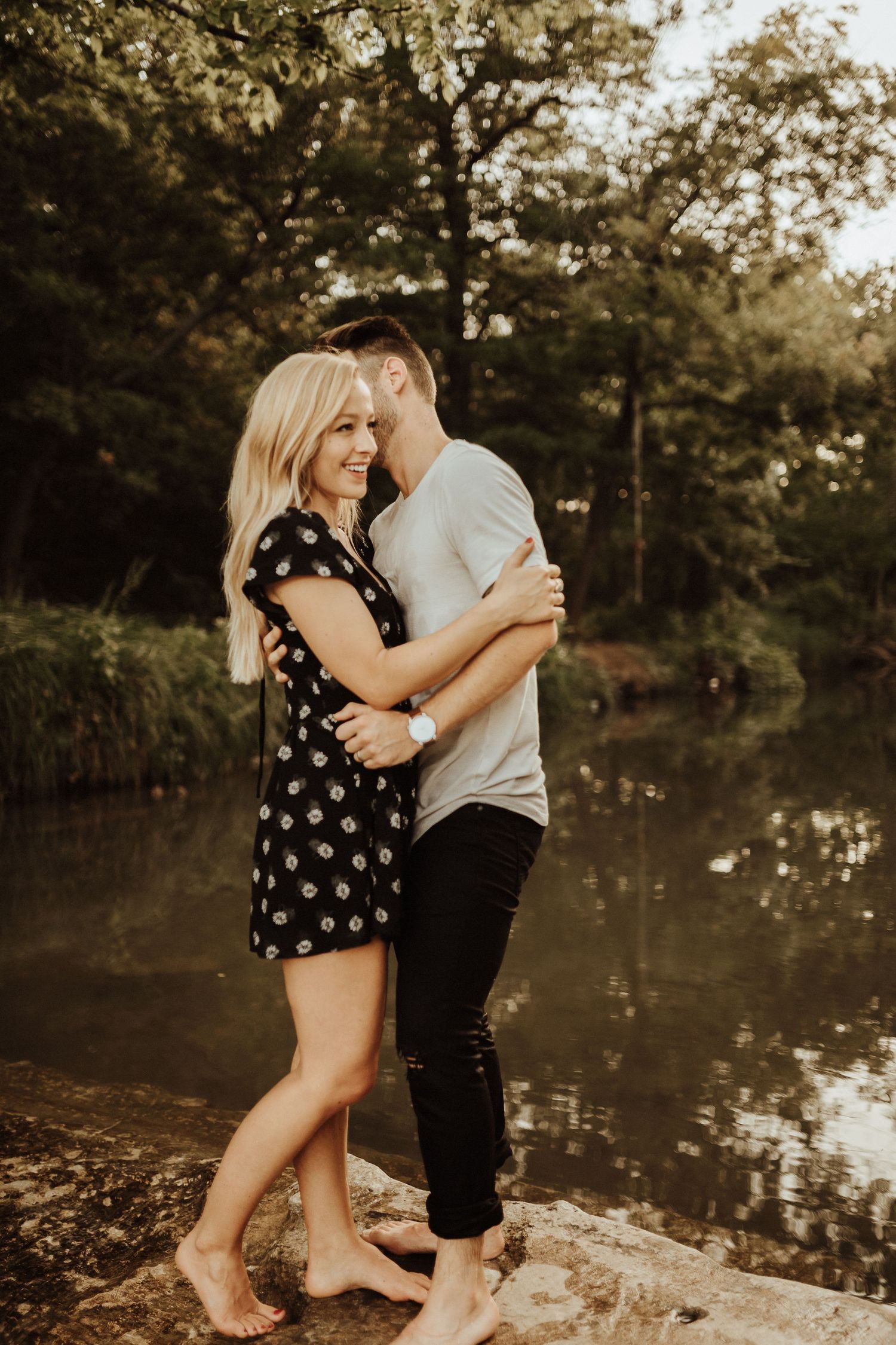 A couple embraces on the edge of a serene pond during golden hour in a wooded area.