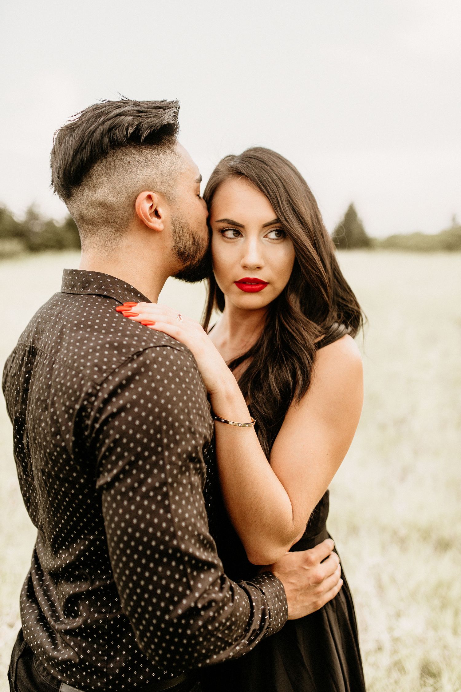 A romantic couple embraces in a sunlit field, she wears a black dress and red lipstick while he dons a polka dot shirt.