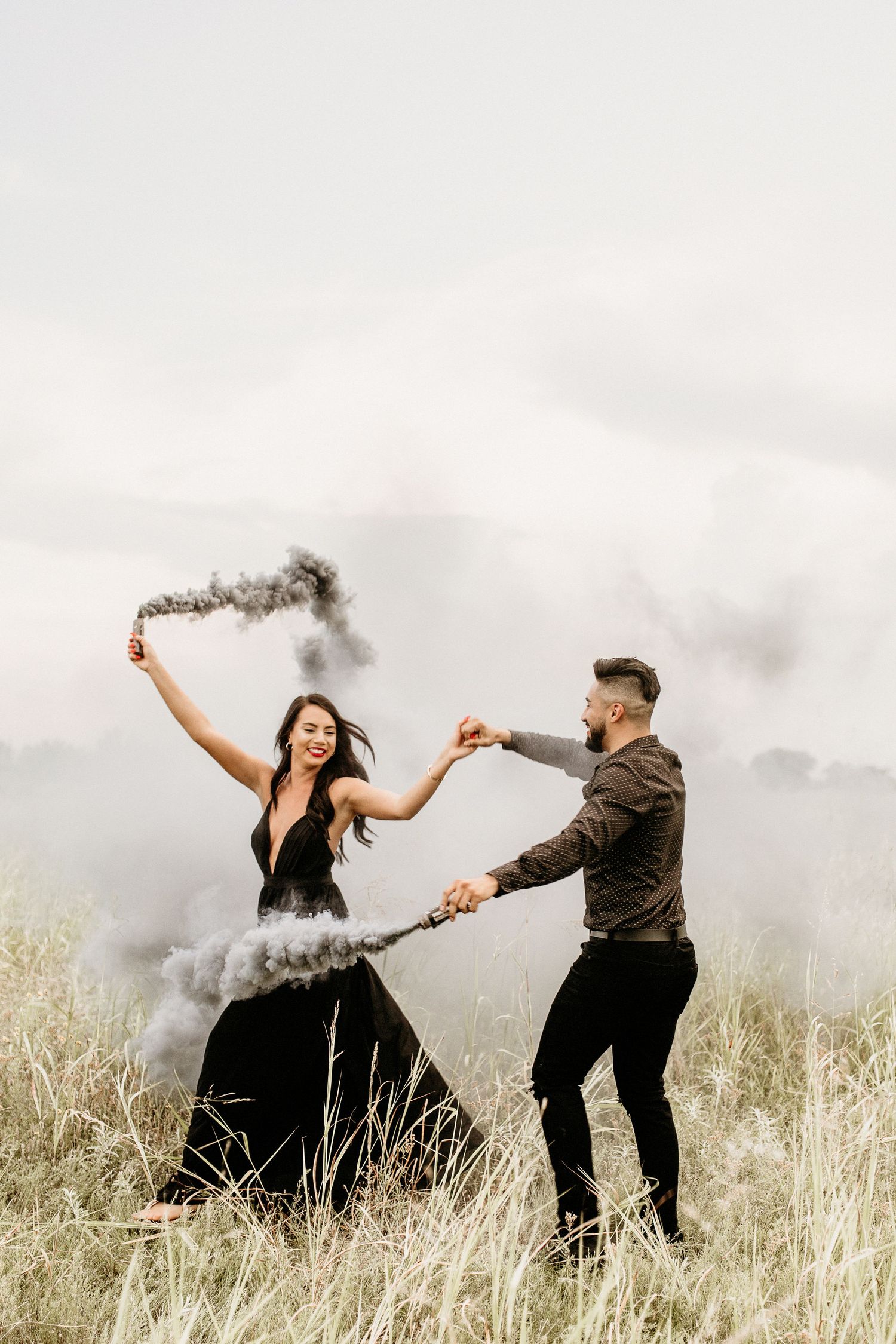 Couple in formal attire hold smoke bombs while dancing in a grassy field on cloudy day.
