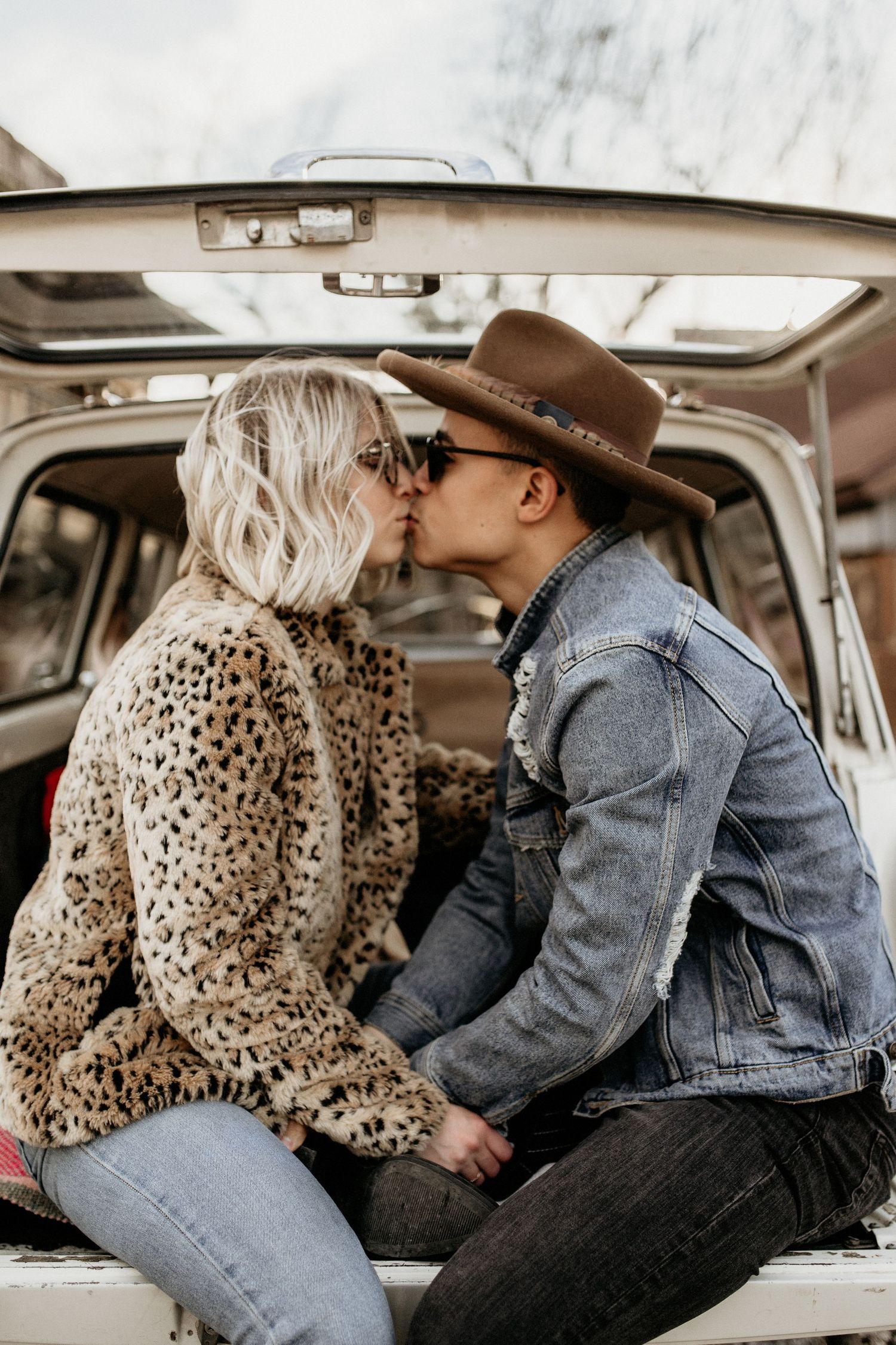 Couple sharing a romantic kiss in the back of a van, wearing a leopard print sweater and denim jacket with brown hat.
