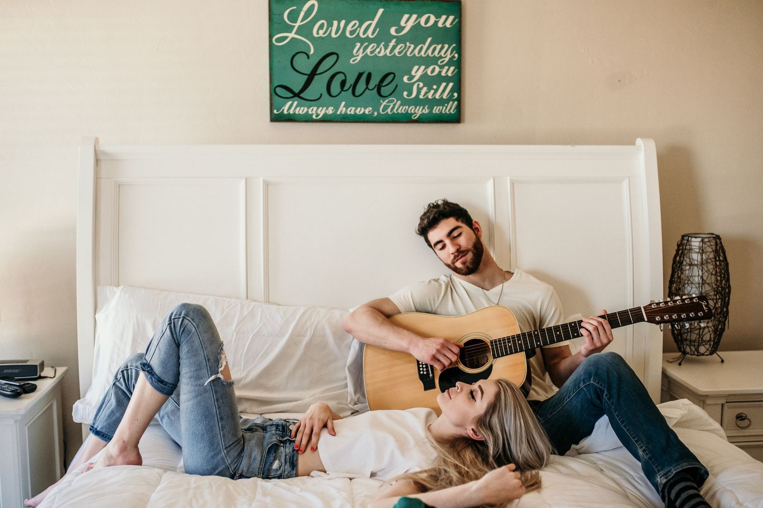 A person sits on a cozy white bed while playing acoustic guitar with a decorative sign on the wall.