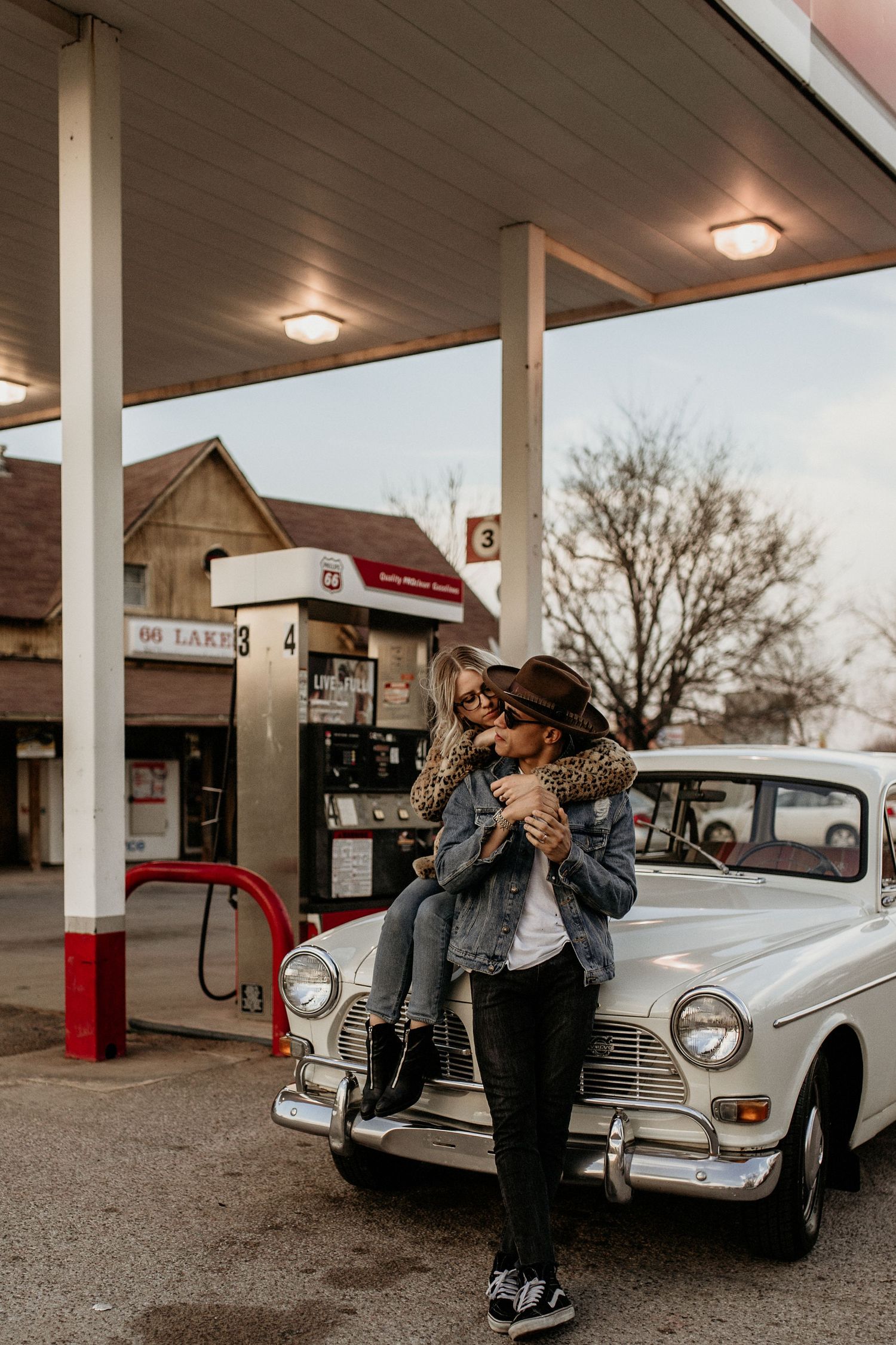 Retro vintage car at old gas station with figure in denim jacket and hat leaning against white classic automobile.