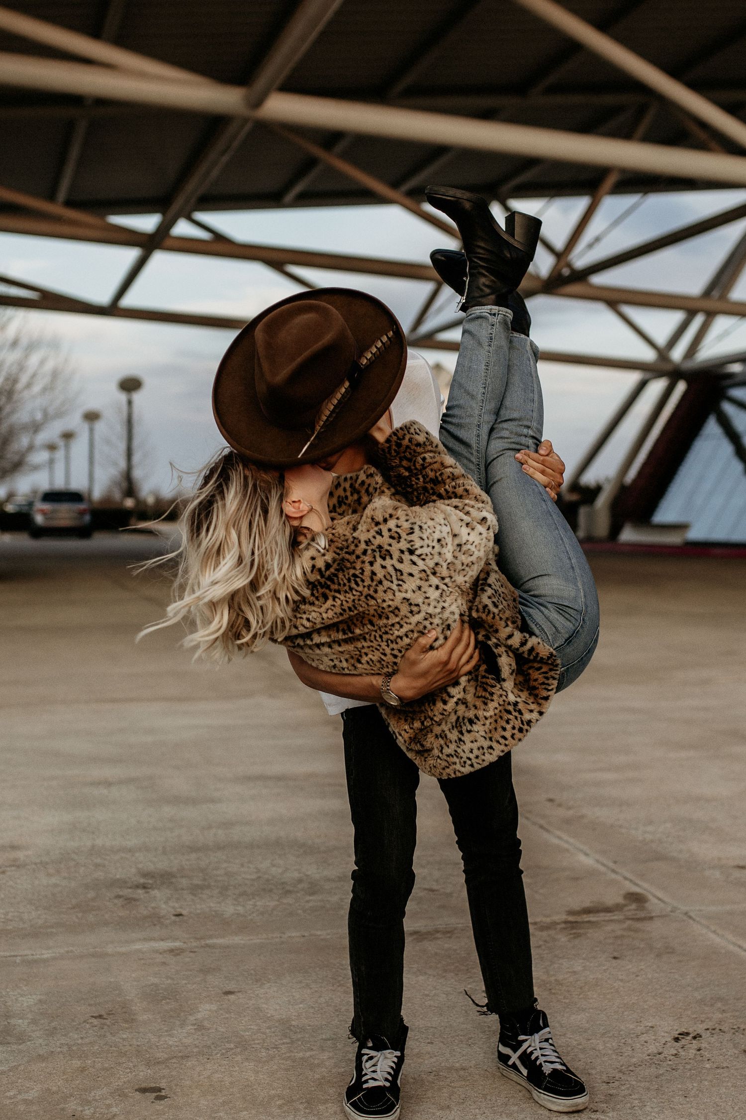 Woman in leopard print jacket and brown hat playfully dipping backwards in covered urban area.