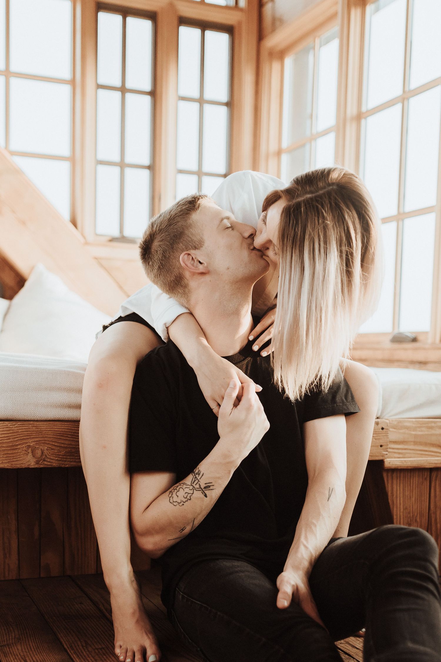 A romantic couple in black outfits share an intimate moment by bright windows in warm natural lighting.