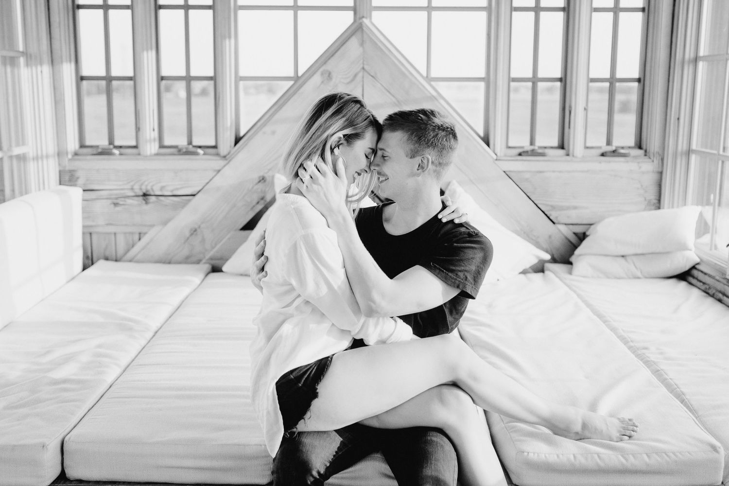 Black and white romantic photograph of couple embracing on bed with white sheets and pillows in sunlit room.