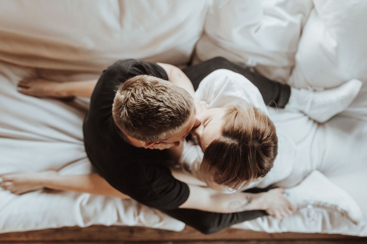 High angle view of two people lying on white bedding, embracing in intimate moment from above.