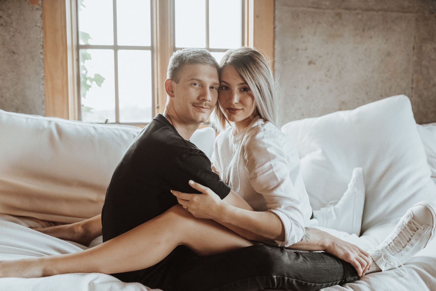 A couple cuddles together on a white couch near a bright window in a cozy indoor setting.