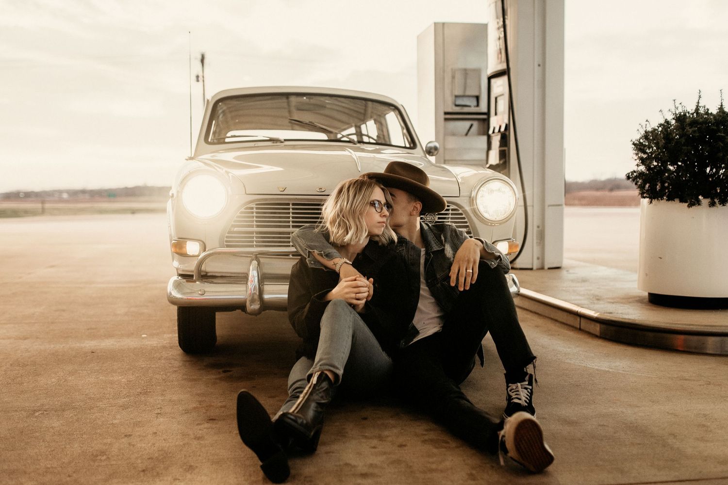 A couple sits on the ground in front of a vintage car at a retro gas station in sepia tones.