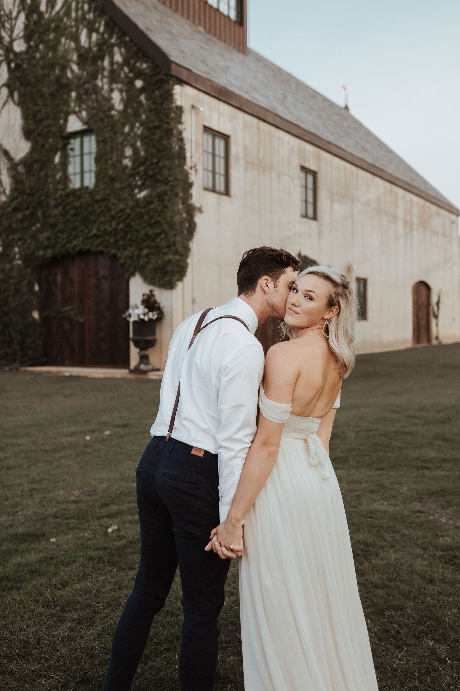 A couple in wedding attire share an intimate moment outside a rustic barn with ivy-covered walls.