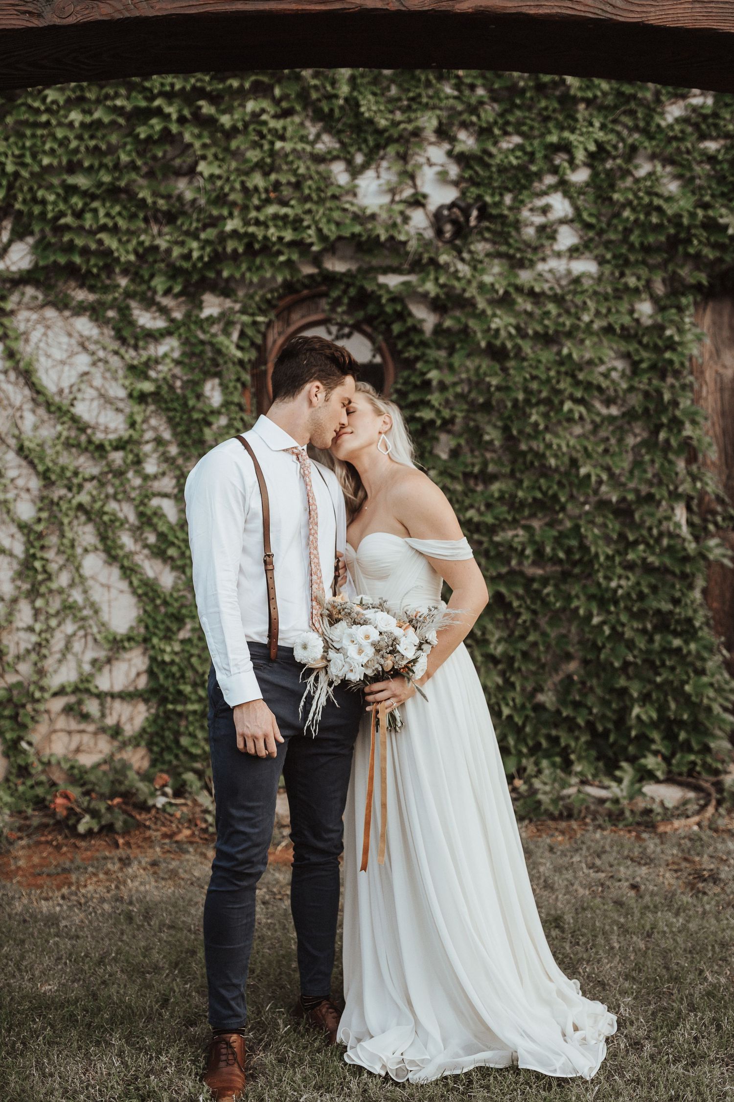 A couple shares an intimate moment in front of an ivy-covered wall, with the bride in white and groom in suspenders.