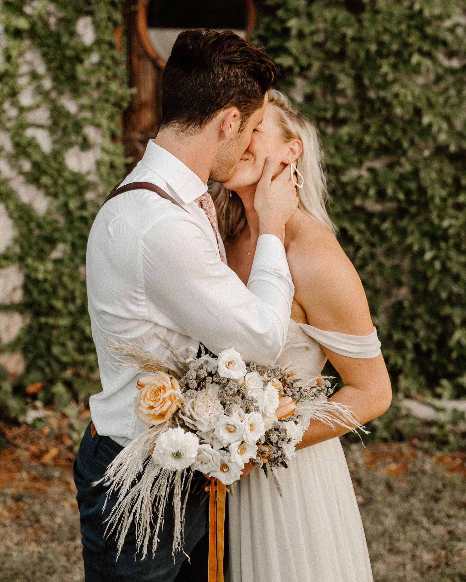 Couple shares intimate moment at rustic outdoor wedding with white and peach floral bouquet with pampas grass details.