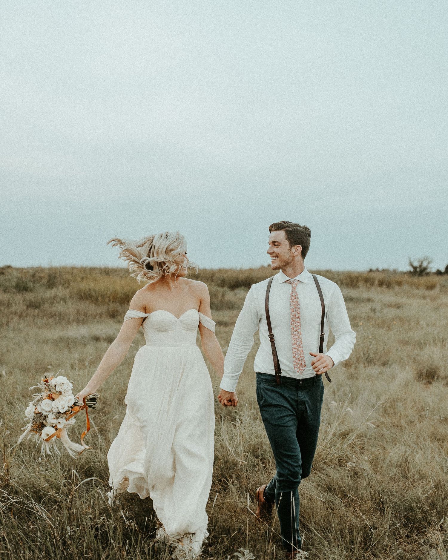 A couple walks through a grassy field, with the bride in a flowing white dress and groom wearing suspenders.