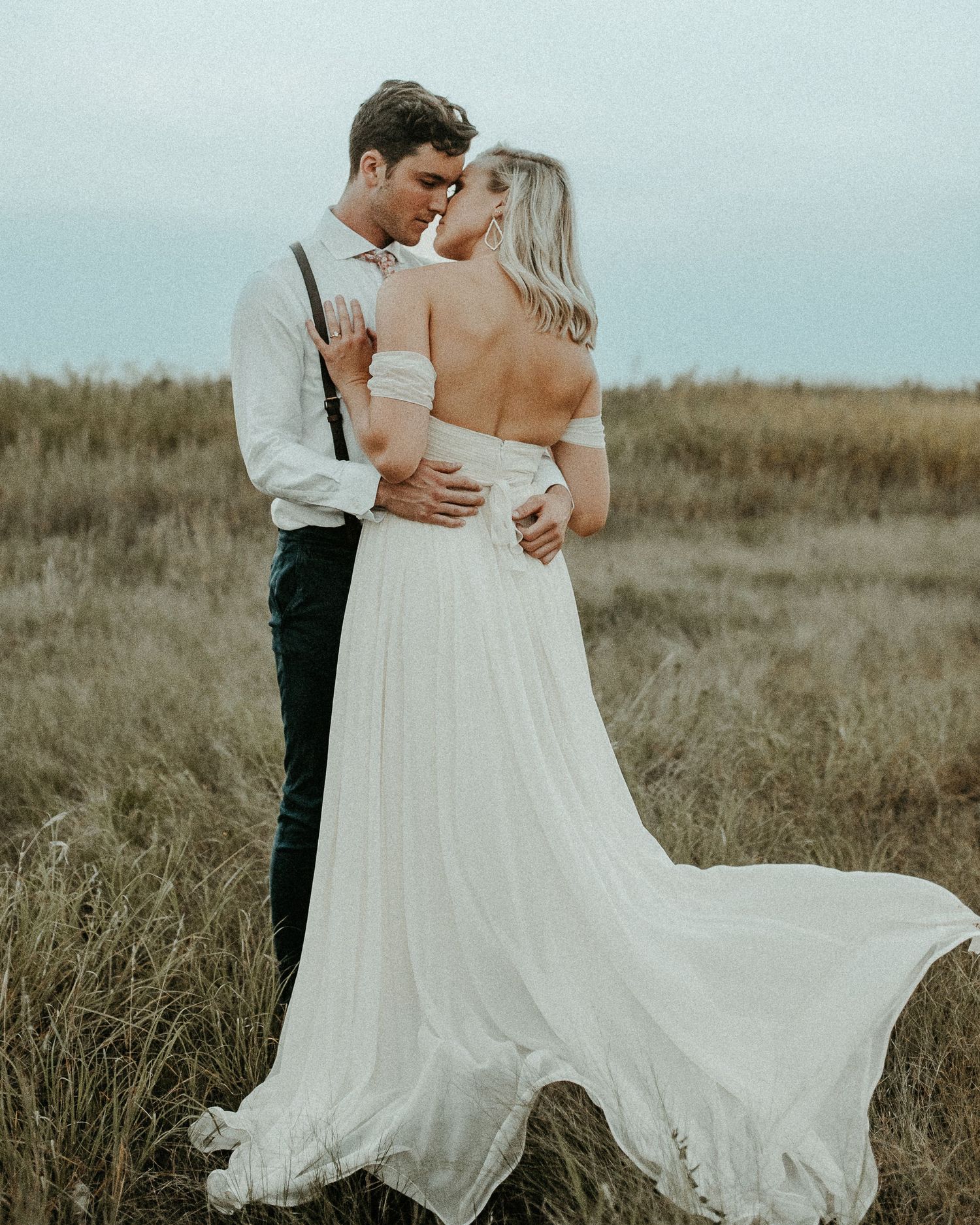 A couple in wedding attire embrace in a field with a flowing white dress catching the breeze at sunset.