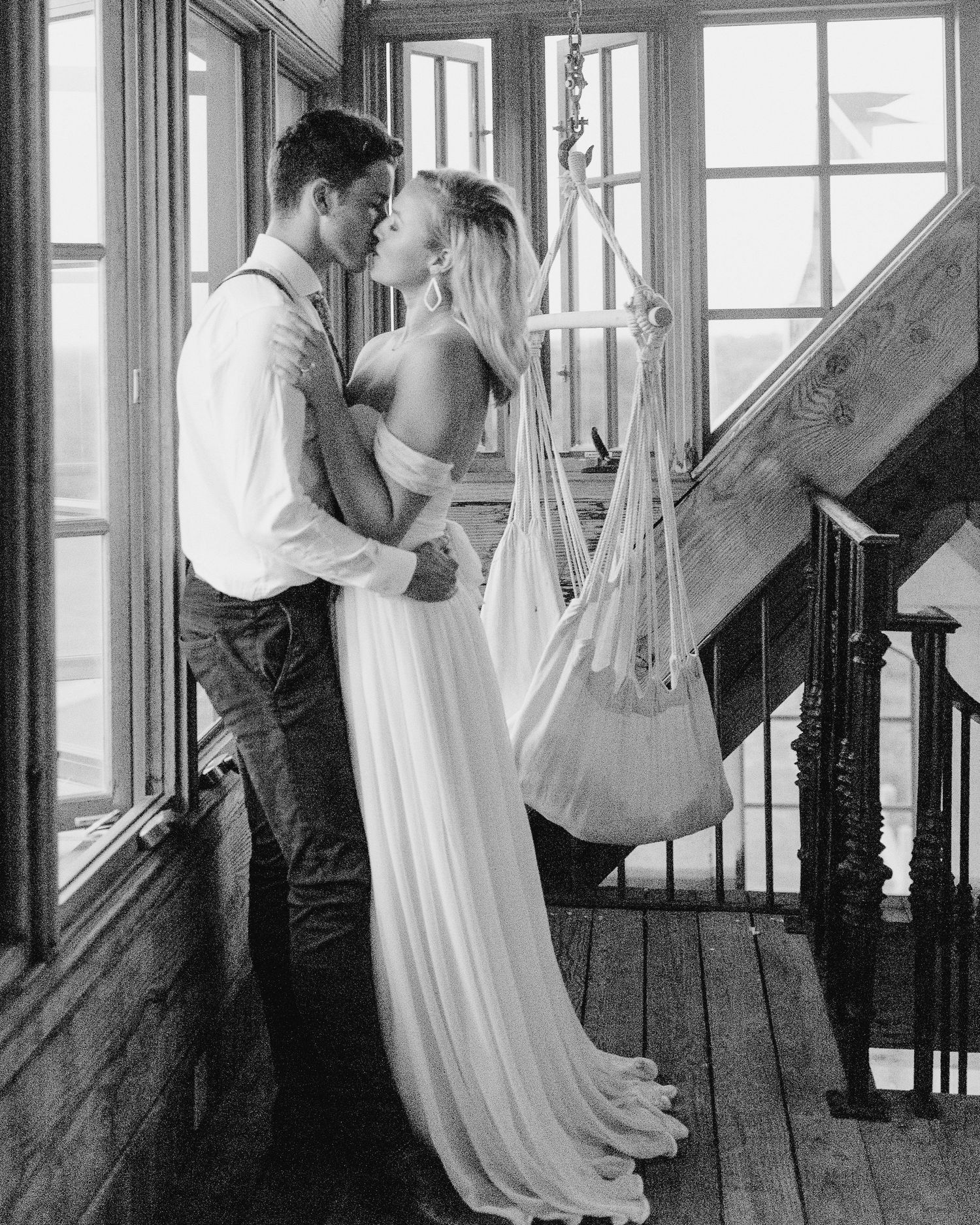A romantic black and white portrait of a couple embracing in a sun-lit stairwell with flowing white dress and vintage decor.